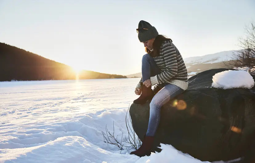 Icebug boot lifestyle image of a woman putting on the boots in front of a snowy lake- kumpfy shoes